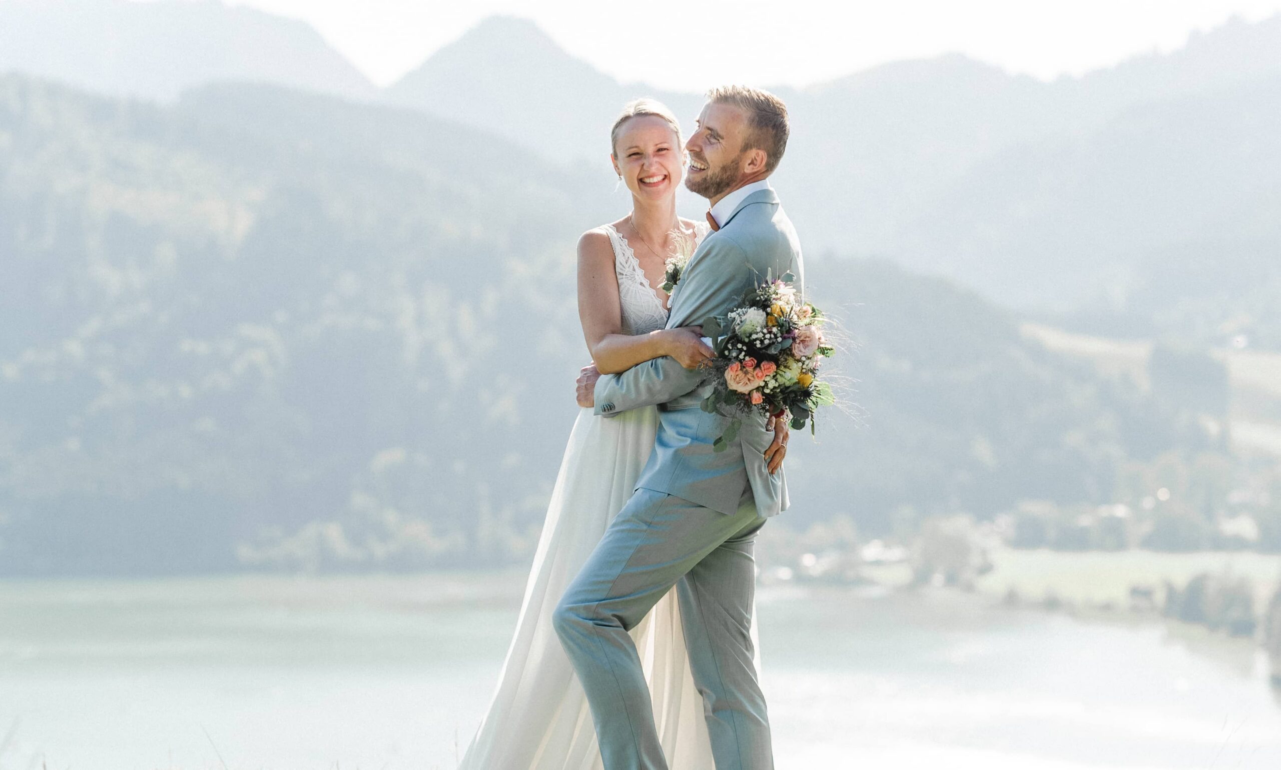 Groom lifting the bride by a lake with mountains in the background