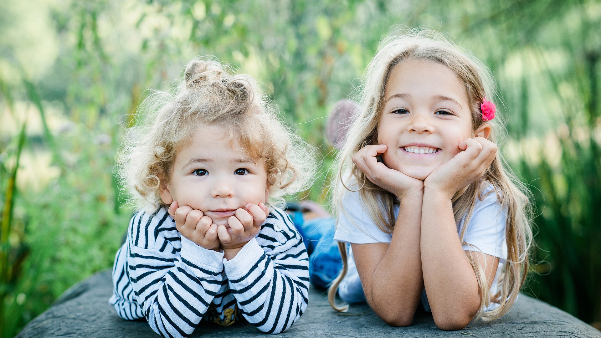Two children outdoors, smiling with their chins resting on their hands