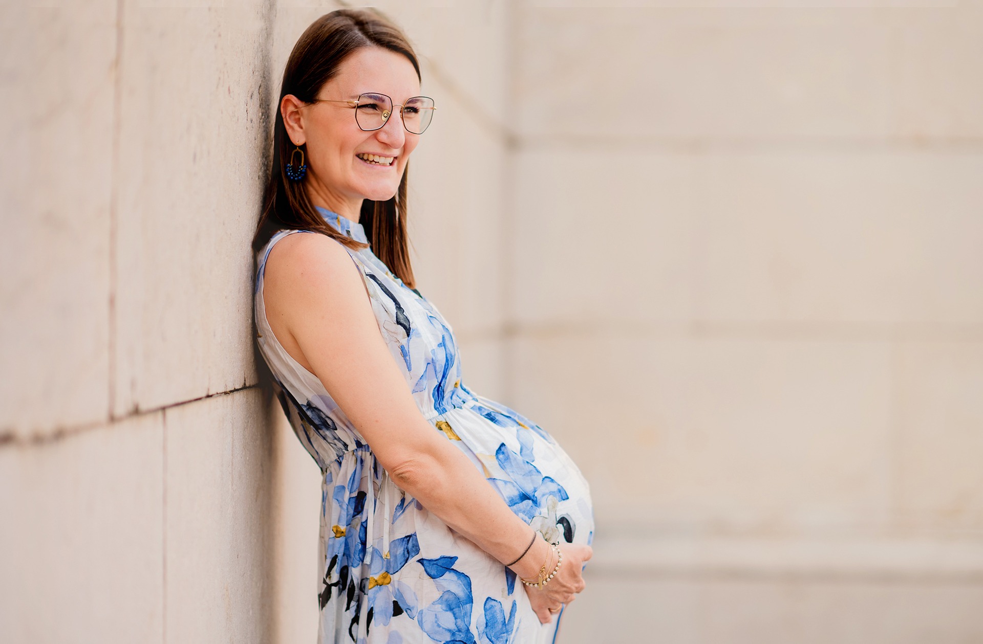 Expectant mother in a floral dress leaning against a pale stone wall