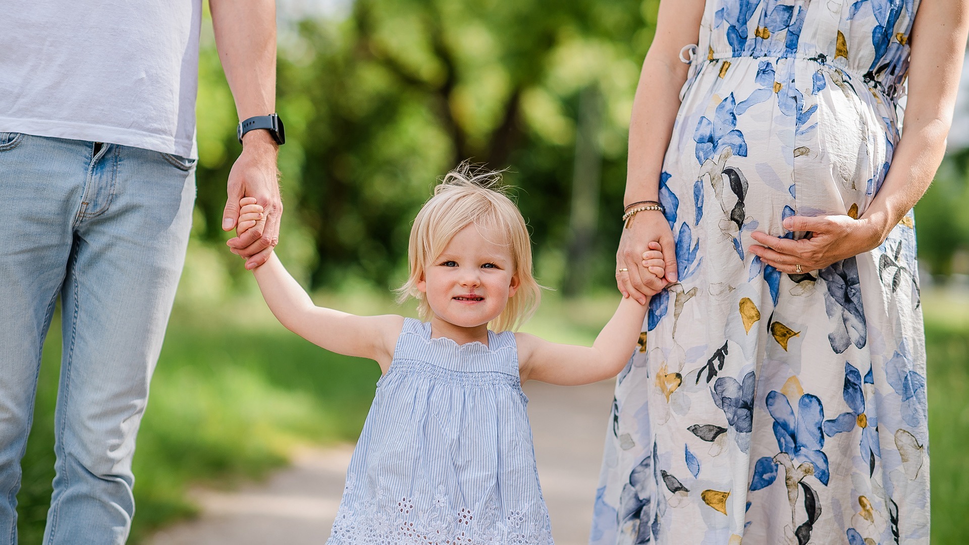 Kind hält draußen die Hände seiner Eltern neben dem Babybauch der Mutter