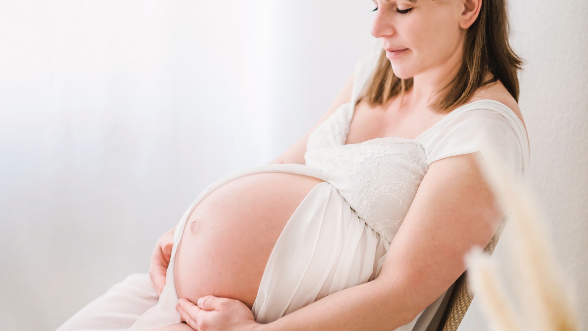 Sitzende Schwangere in einem weißen Kleid mit den Händen am Bauch